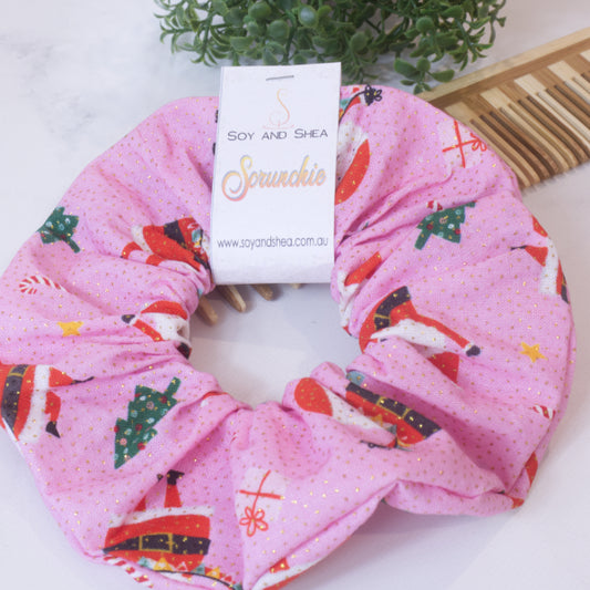 A colorful scrunchie sits on wooden surface resting against a wooden comb with greenery in the background.  A cardboard tag reads SOY AND SHEA SCRUNCHIE.    The scrunchie is bright pink with sparkling gold dots and features an images of santa, presents, candy canes and Christmas trees randomly placed.