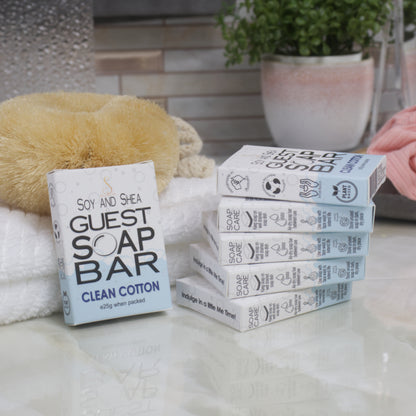 To the left, a box of Clean Cotton Guest Soap bar rests against a while towel with body brush.  The right is a stack of 5 other boxes.  