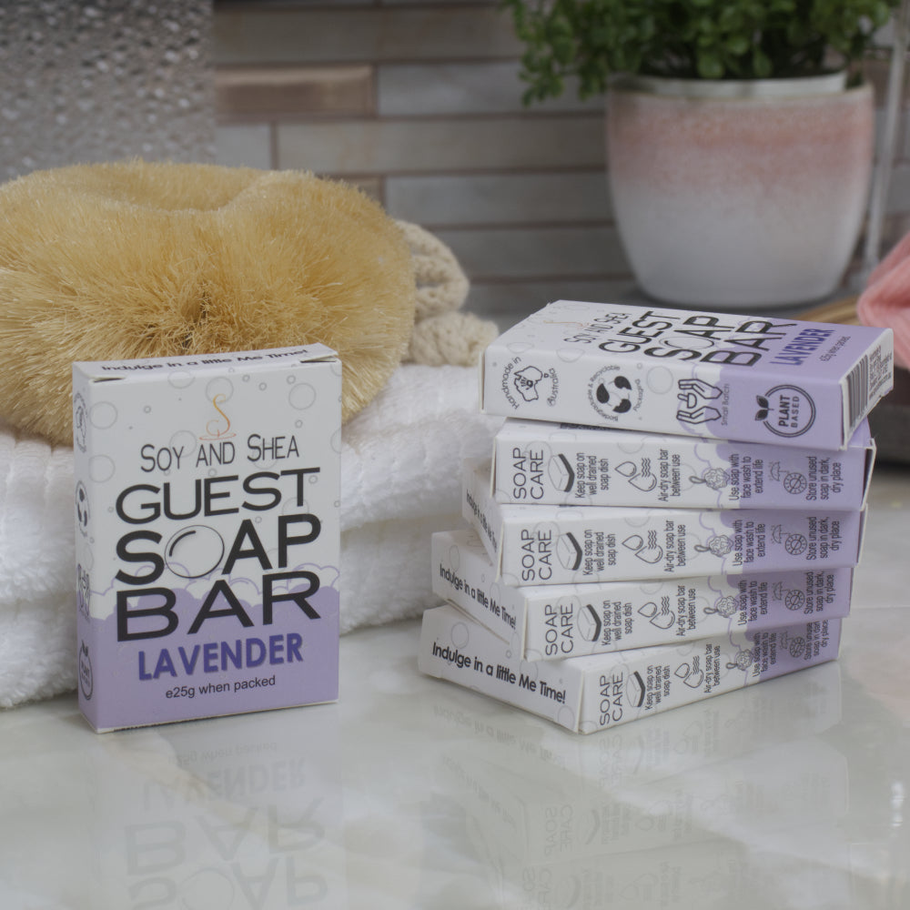To the left, a box of Lavender Guest Soap bar rests against a while towel with body brush.  The right is a stack of 5 other boxes.  