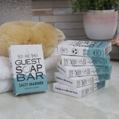 To the left, a box of Salty Mariner Guest Soap bar rests against a while towel with body brush.  The right is a stack of 5 other boxes.  