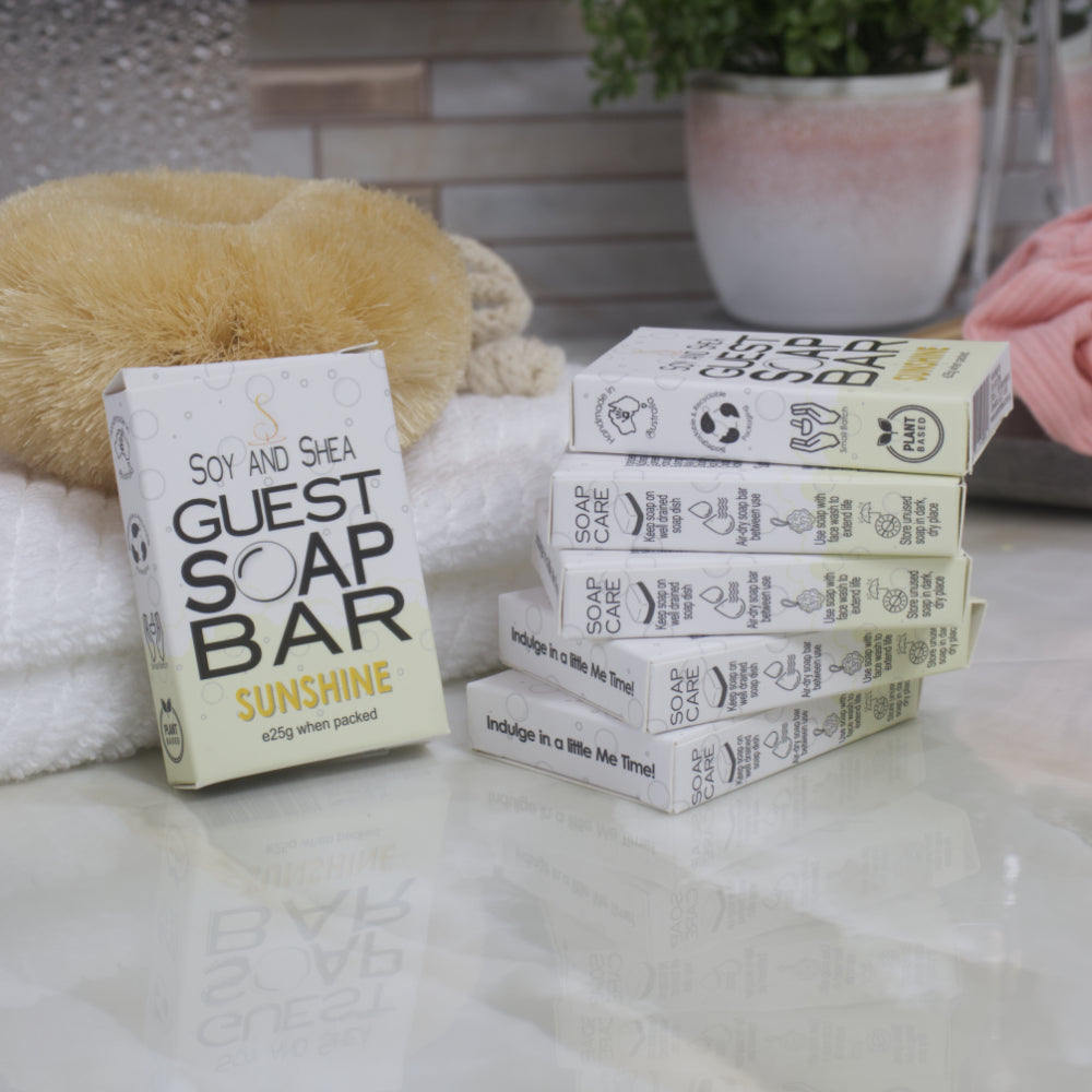 To the left, a box of Sunshine Guest Soap bar rests against a while towel with body brush.  The right is a stack of 5 other boxes.  