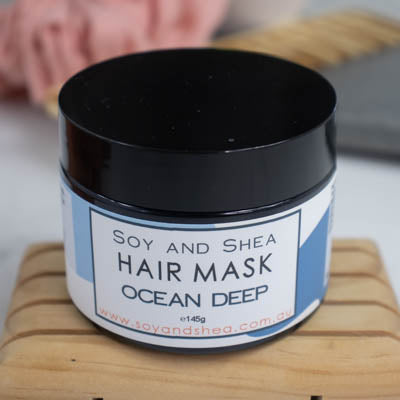 A black jar with lid on sits on a wooden surface against a blurred background with scrunchie and comb. The jar features a white label with a blue abstract design with text HAIR MASK OCEAN DEEP.