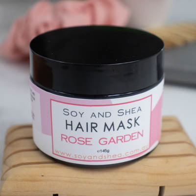 A black jar with lid on sits on a wooden surface against a blurred background with scrunchie and comb. The jar features a white label with a pink abstract design with text HAIR MASK ROSE GARDEN.