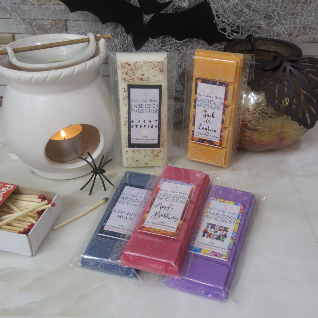 A collection of Soy Wax Snap Bars in various colors (blue, red, purple, yellow, and white) with spooky themed labels, arranged alongside a white ceramic fragrance warmer with a lit tea light, a box of matches, and Halloween decorations on a marble surface.