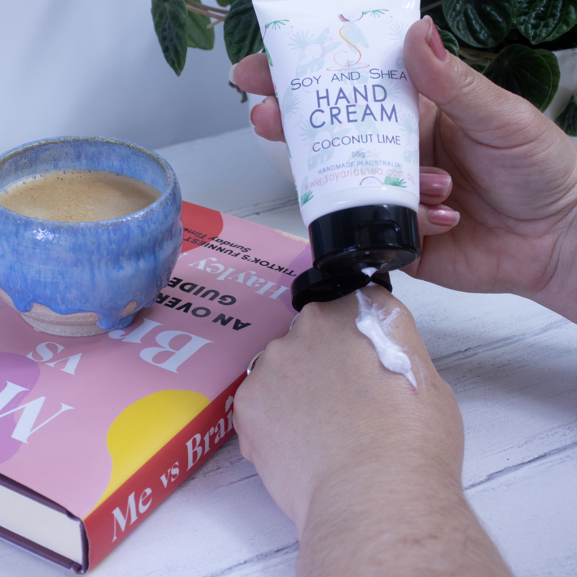 A person is squeezing Soy and Shea's Coconut Lime Hand Cream, infused with nourishing camellia oil, onto their hand from the tube. A blue ceramic cup and a book titled "Me vs Brain" rest on a white table nearby, with green leaves of a plant visible in the background.