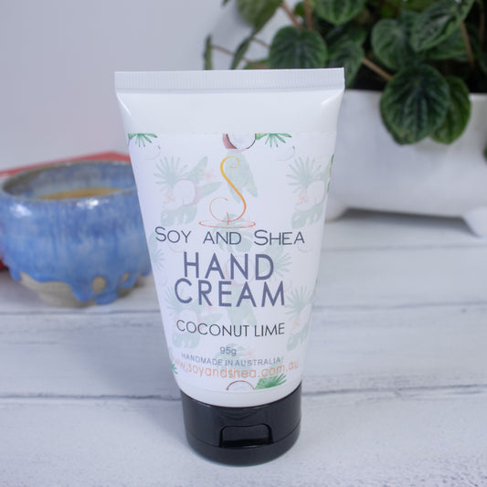 A 95g tube of Soy and Shea's Coconut Lime Hand Cream is displayed on a light surface. The tube, enriched with hemp seed oil, has a white background with a tropical design featuring coconuts and palm leaves. In the background, there are blurred plants and a ceramic pot.