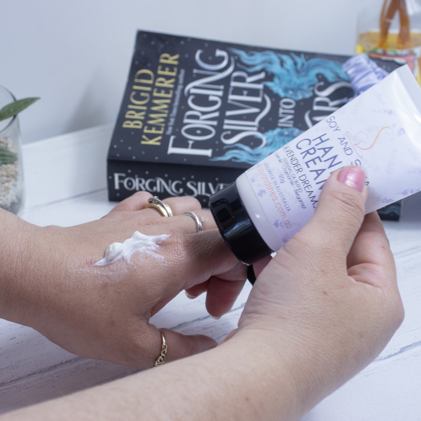 A person applies deeply moisturizing hand cream from a tube labeled "Lavender Dreams Hand Cream (Essential Oil Blend)" by Soy and Shea onto their hand. In the background a copy of the book "Forging Silver into Stars" by Brigid Kemmerer is visible on a white surface along with a small plant.