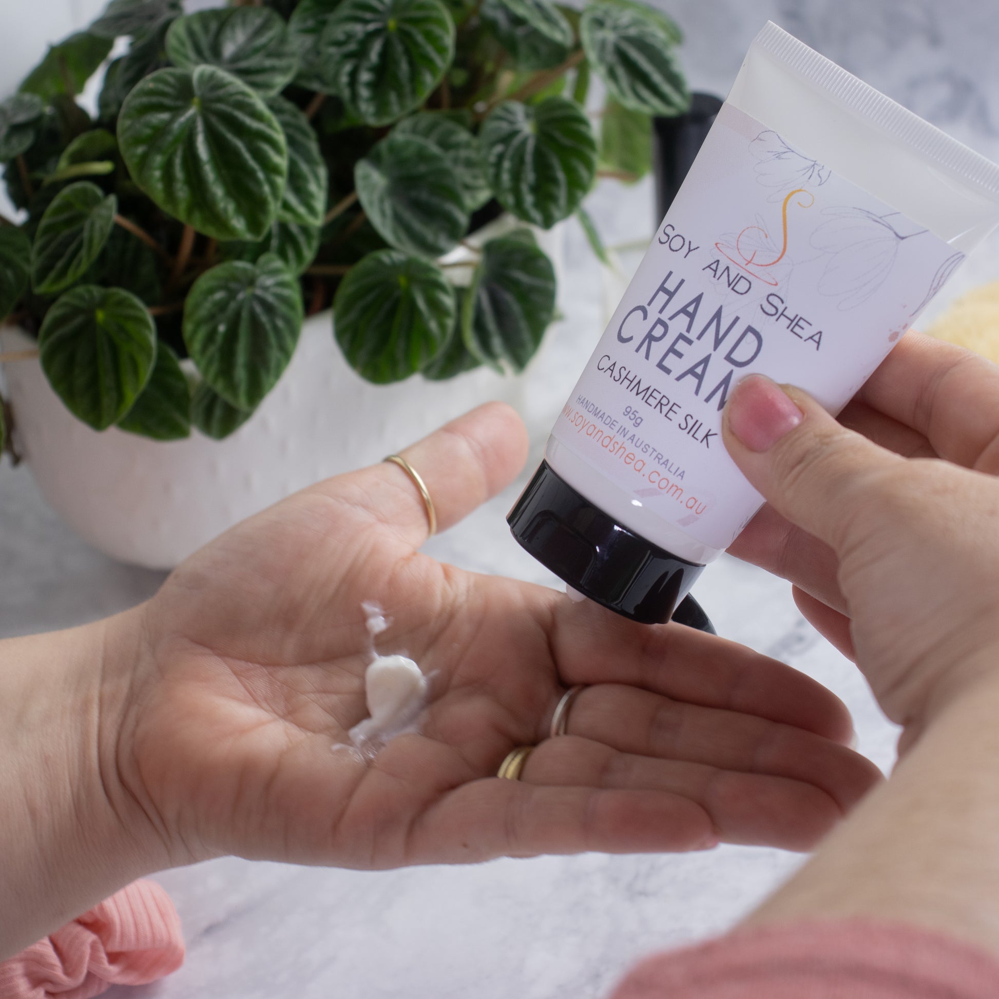 A person is squeezing Soy and Shea's Cashmere Silk Hand Cream from a white tube with black lid onto their open palm. In the background, there is a potted green plant with heart-shaped leaves. The scene is set on a light-colored surface.