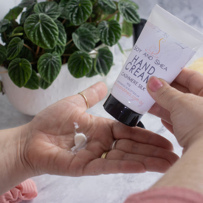 A person is squeezing Soy and Shea's Cashmere Silk Hand Cream from a white tube with black lid onto their open palm. In the background, there is a potted green plant with heart-shaped leaves. The scene is set on a light-colored surface.