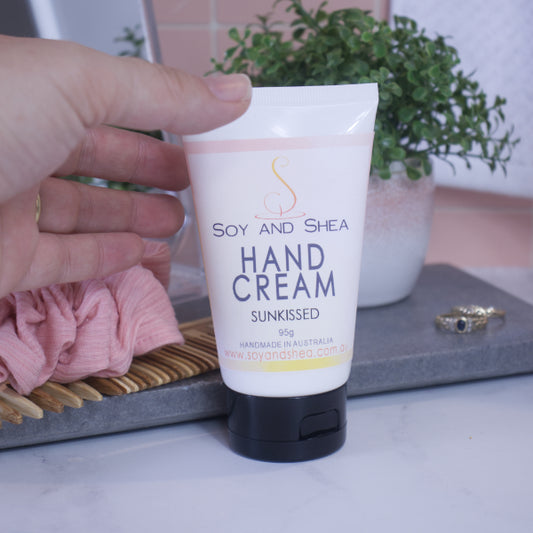 A hand reaches to pick up a white tube with black lid of Hand Cream which is sitting on a bathroom counter top with a plant, gold rings and a pink scrunchie