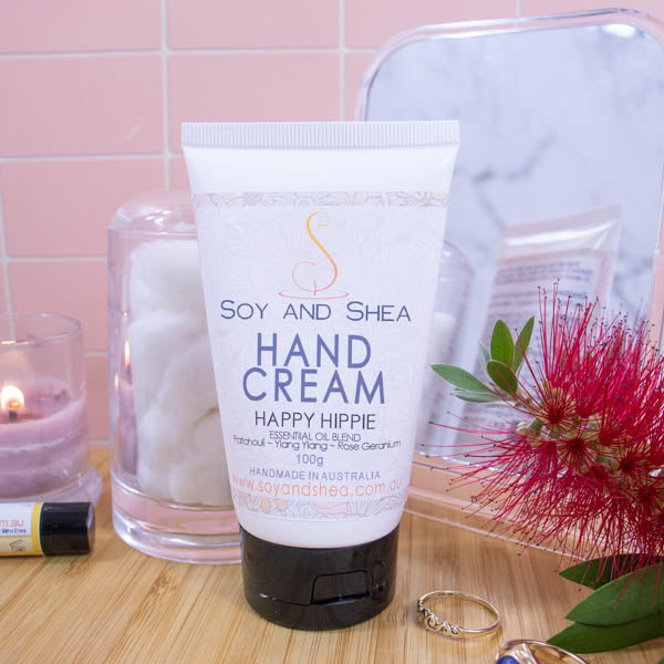 A white tube with black flip-top lids stands on a wooden surface with a pale pink tile background.  To one side is a burning candle and jar of cotton balls and on the is a red bottle brush flower and some gold rings. Behind is a mirror reflecting the back of the tube.