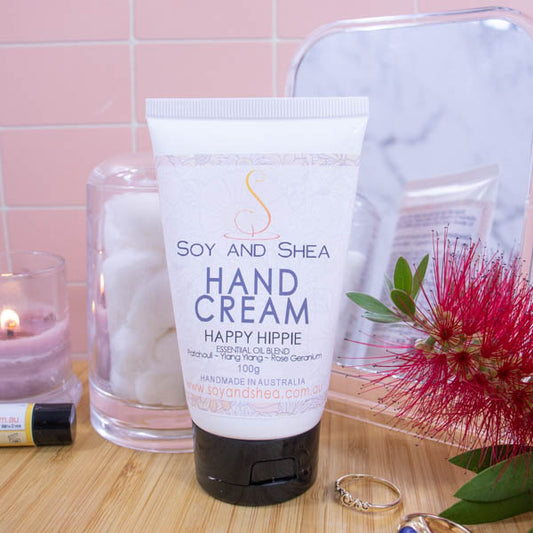 A white tube with black flip-top lids stands on a wooden surface with a pale pink tile background.  To one side is a burning candle and jar of cotton balls and on the is a red bottle brush flower and some gold rings. Behind is a mirror reflecting the back of the tube.