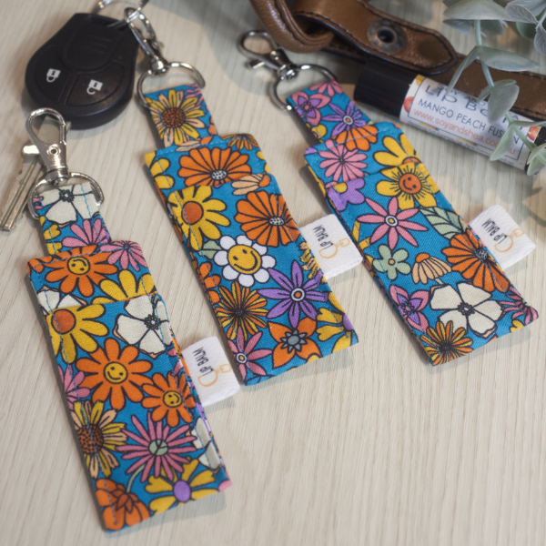A selection of three clip balms showing variations in fabric design, lie on a wooden side with the middle Clip Balm attached to a car key.  A lip balm lays near-by along with some greenery.  The Clip balms have a bright blue background with a variety of flowers in yellow, orange, pink and white with the occassional flower showing a smiling face