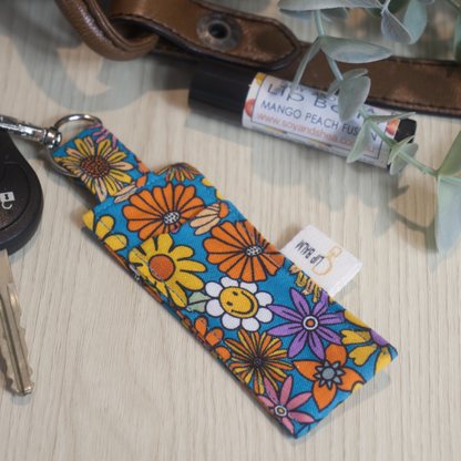 A clip balm holder lies on a wooden surface attached to a car key with a "lobster claw" clip.  To the side is a tube of lip balm surrounded by greenery and a leather strap from a bag.  The Clip Balm has a bright blue background and has drawings of brightly coloured flowers in various styles and colours including white, purple, orange and yellow.  Some flowers feature a smiling face.