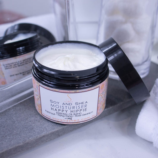 A black jar with a colourful floral label sits on a stone tray with the lid resting on the side revealing a white cream.  The scene is reflected in a mirror which stads next to a jar of cotton balls.