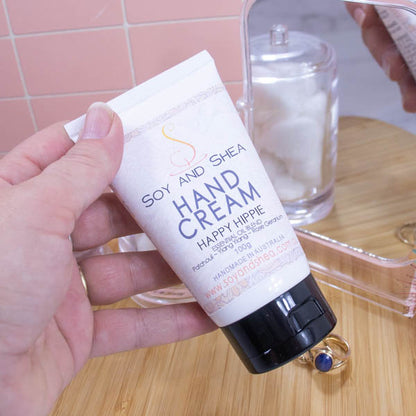 A hand holds a white tube with black lid labelled HAND CREAM against a background of a wooden surface with pink tile wall.  A mirro to the right shows the reflection.