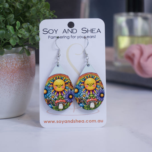 A pair of earrings on silver hooks hang on a white card branded with SOY AND SHEA leaning against a pot with a green leafy plant and a blurred background.  The earrings are teardrop shaped with a cartoon drawing of a large sun with kawai face in front of a rainbow.  Below is a toadstool with a green frog surrounded by bright flowers and grass