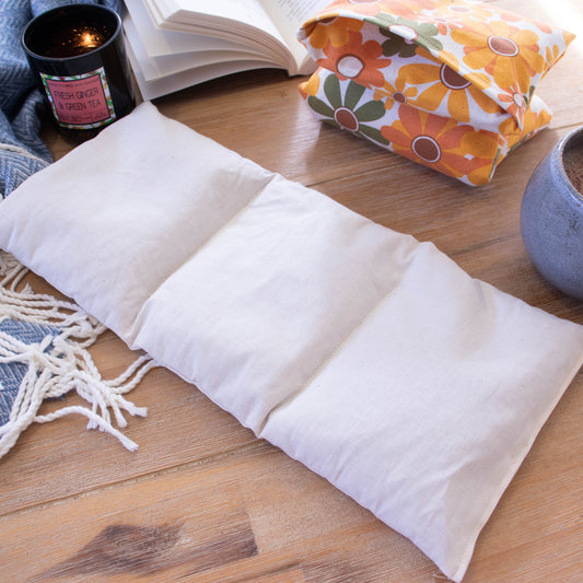 The insert of a Heat Pack lays on a wooden surface next to a burning candle, open book and filled coffee cup.  To the right is a folded heat pack.  The insert is a long rectangular shape made of  a cream fabric.  It has two lines of stitching going from long side to long side dividing the insert into 3 equal parts.