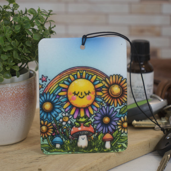 A car air freshener rests against a potted plant with a cartoon image of a smiling sun above a toadstool with a frog all surrounded by flowers and a rainbow.  Next to the air freshener is a bottle of scent and some car keys