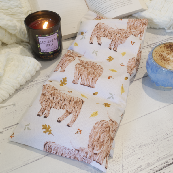 A rectangular heat pack with a pattern of illustrations of highland cows, leaves and autumn-themed designs on white fabric, displayed on a wooden surface alongside a burning candle and a cup of coffee.