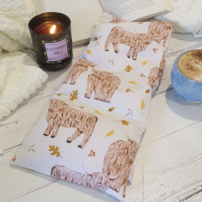 A rectangular heat pack with a pattern of illustrations of highland cows, leaves and autumn-themed designs on white fabric, displayed on a wooden surface alongside a burning candle and a cup of coffee.