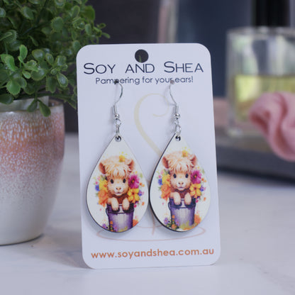 A pair of earrings on silver hooks hang on a white card branded with SOY AND SHEA leaning against a pot with a green leafy plant and a blurred background.  The earrings are teardrop shaped featuring a baby cow with a fluffy head sitting in grey bucket surround by flowers