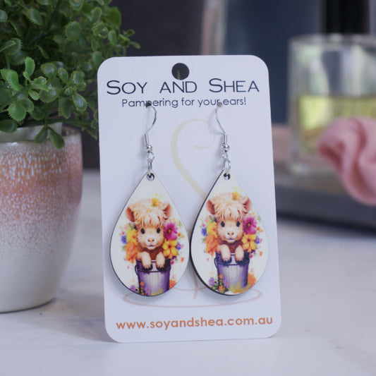 A pair of earrings on silver hooks hang on a white card branded with SOY AND SHEA leaning against a pot with a green leafy plant and a blurred background.  The earrings are teardrop shaped featuring a baby cow with a fluffy head sitting in grey bucket surround by flowers