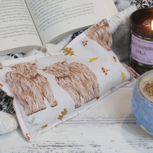 A Soy and Shea Highland Cows Mini Heat Pack with illustrated highland cows and leaves sits on a cozy blanket beside an open book, purple candle, blue mug with a frothy drink.