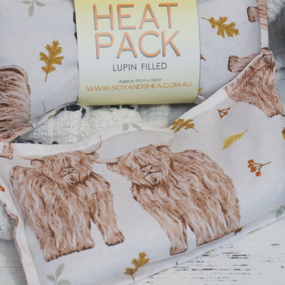 A close up shot of the mini Highland Cows Heat Pack by Soy and Shea shows a white pack with miniture Highland Cows surround by autumn folliage like oak leaves.  A second pack appears in the background with a simple band around the center reading HEAT PACK LUPIN FILLED