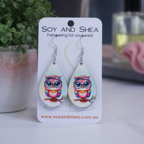 A pair of earrings on silver hooks hang on a white card branded with SOY AND SHEA leaning against a pot with a green leafy plant and a blurred background.  The earrings are teardrop shaped with an illustration of a pink owl with rainbow wings and bandana around head standing on a tree branch against a blue background