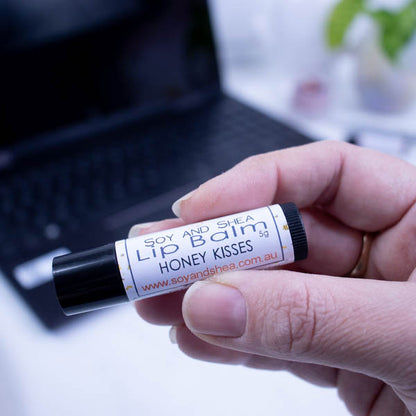 A hand holds a black tube of lip balm against a blurred background with an open laptop computer on a white surface.  The label has text LIP BALM HONEY KISSES on a white background with a border of bees in flight.