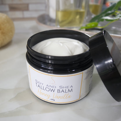 A black jar with a decorative label is displayed on a bathroom countertop.  The label identifies it as Tallow Balm in Honey Vanilla Scent.  The lid of the jar rests on the side of the jar revealing a white balm inside.