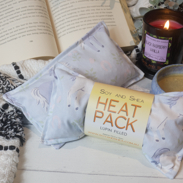 Two Mini Heat Packs rest on a wooden table top resting against an open book surround by a black and white blanket, burning candle and filled coffee cup.   The heat packs are a light blue colours with illustrations of white and blue horses.  The Heat Pack towards the front has a belly band label reading HEAT PACK LUPIN FILLED