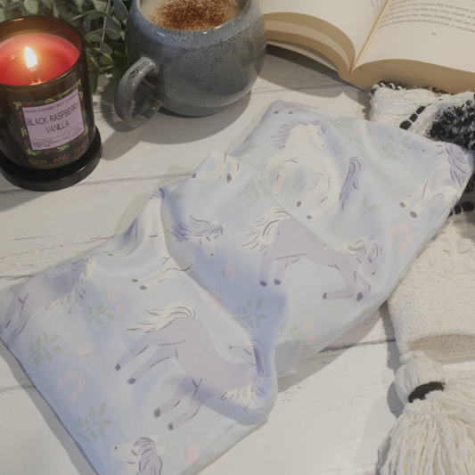A small rectanglular Heat Pack rests on a wooden table top along with a black and white blanket, open book, burning candle and filled coffee cup evoking a cosy scene.  The Heat pack is a pale blue material with illustrations of hores in white and pale purple wiht sprigs of leaves and pinks horse-shoes