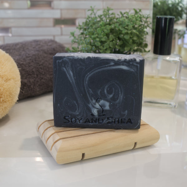 A black soap with white swirls sits on a wooden soap rack on a bathroom counter top along with a grey towel and potted plant against a tiled background.