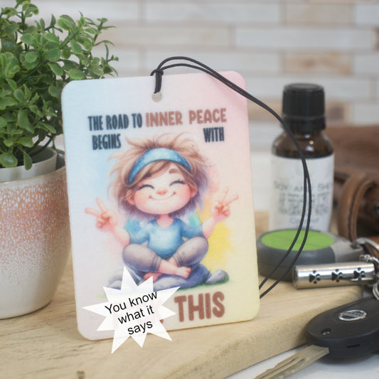 The Inner Peace Car Air Freshener by Soy and Shea features a cartoon lady making peace signs on a hanging card that reads, "The road to inner peace begins with THIS." A word has been blocked out with star