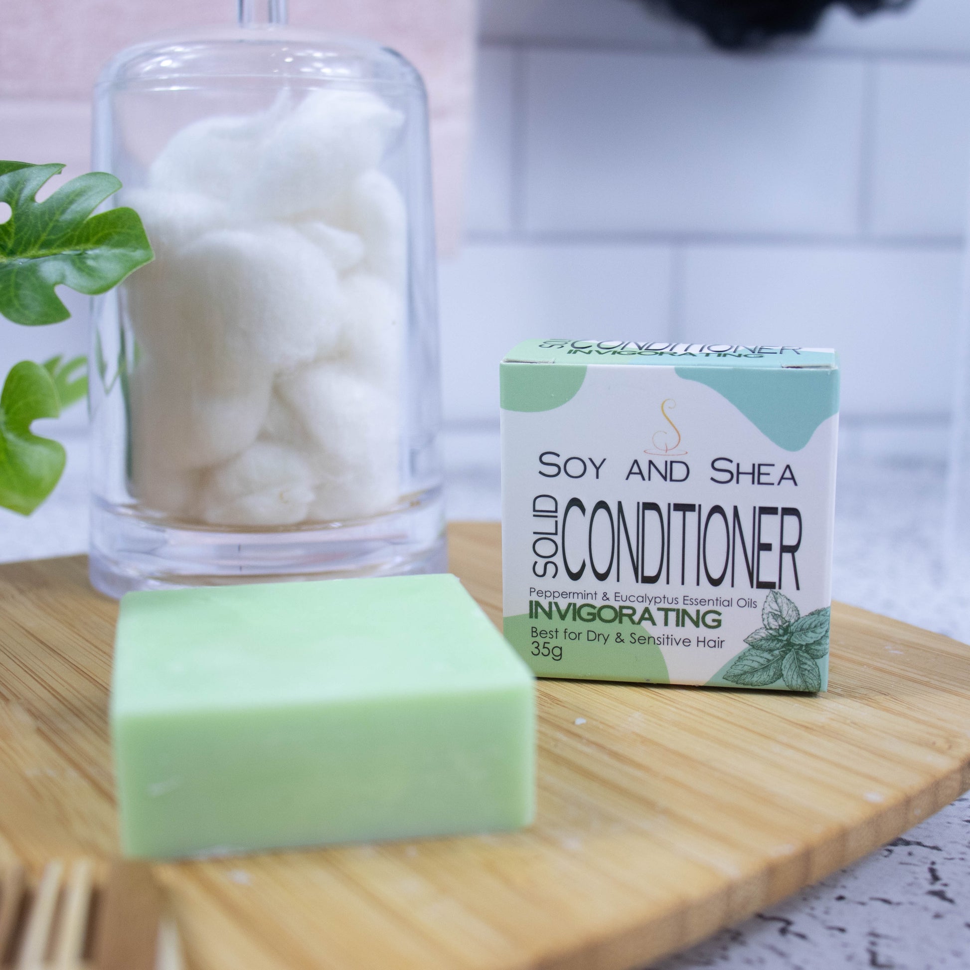 A wooden tray sits on a bathroom counter with a potted plant and jar of cotton balls against a blurred tiled background. Towards the front of the tray stands a white box with green abstract pattern with text SOLID CONDITIONER and next to it stands a square pale green bar of conditioner.