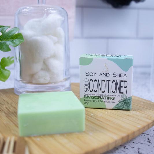 A wooden tray sits on a bathroom counter with a potted plant and jar of cotton balls against a blurred tiled background. Towards the front of the tray stands a white box with green abstract pattern with text SOLID CONDITIONER and next to it stands a square pale green bar of conditioner.