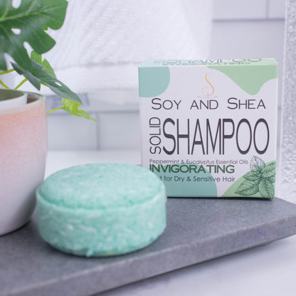 A white box with green abstract pattern and the text SOLID SHAMPOO INVIGORATING stands upright on a grey stone tray with a blurred bathroom background.  Next to the box sits a round bar of solid shampoo.  The bar is mint green with small white lines throughout.