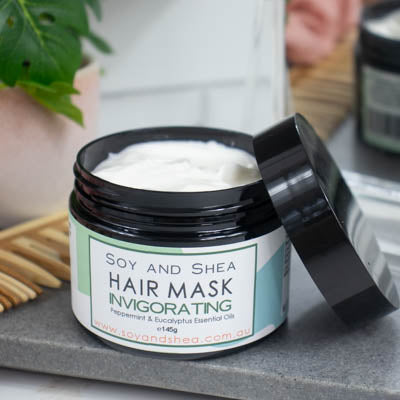 A black jar with the lid resting on its side revealing a white cream inside, sits on a dark grey tile surface against a blurred background of a bathroom counter top. The jar features a white label with greem abstract design with text HAIR MASK INVIGORATING.