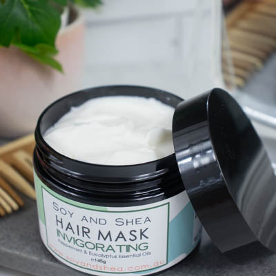 An overhead shot of a black jar with the lid resting on its side revealing a white cream inside, sits on a dark grey tile surface against a blurred background of a bathroom counter top. The jar features a white label with green abstract design with text HAIR MASK INVIGORATING.