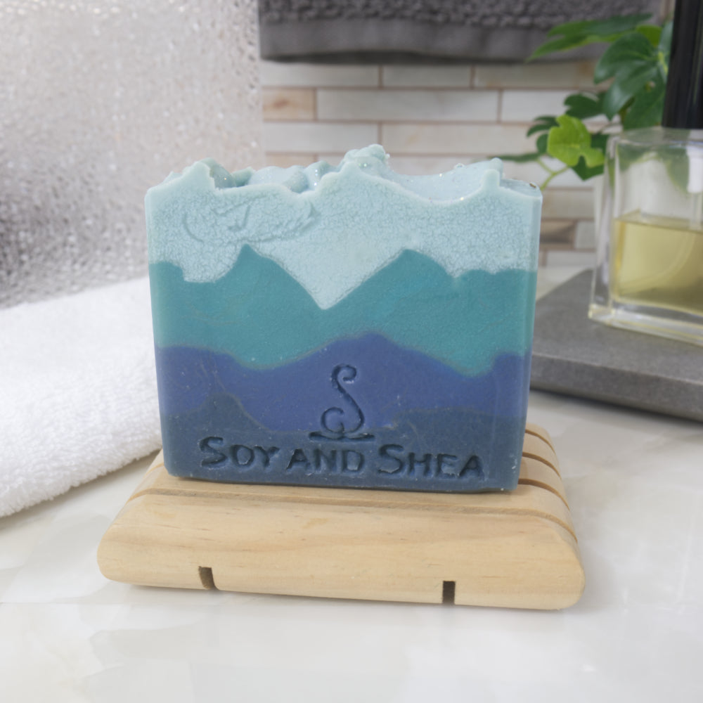 Blue and green layered soap on a wooden stand with 'Soy and Shea' branding with a blurred backgroudn of a bathroom.