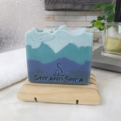 Blue and green layered soap on a wooden stand with 'Soy and Shea' branding with a blurred backgroudn of a bathroom.