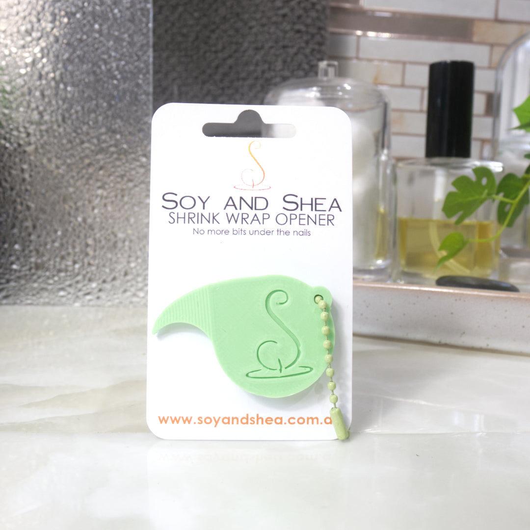 A Jade green Shrinkwrap opener with matching ball and chain keyring is attached to a white card with product details.  The card is standing upright on a bathroom countertop with a bottle of perfume, a jar of cotton balls and towel in the background