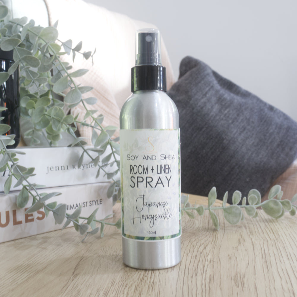 A silver metal spray bottle of 'Soy and Shea Room + Linen Spray' with a black spray nozzle, standing on a light wooden table. The background features a cozy home setting with a beige cushion, a dark gray pillow, stacked books, and green eucalyptus branches.  The label indicates this is Japanese Honeysuckle