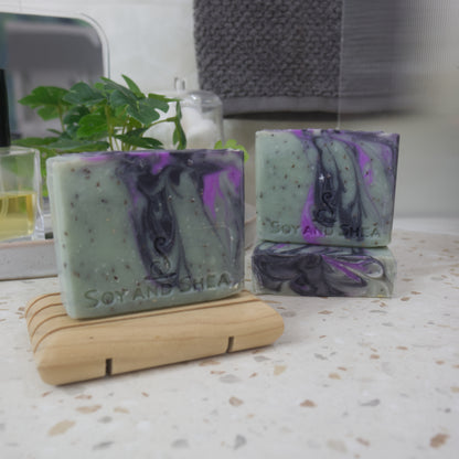 A selection of soaps are displayed on a bathroom countertop with blurred background.  To the front left, a soap bar stands on a wooden soap dish, while to the back right, a soap lays flay on the bench with another standing on it.   The soaps are a forrest green with brown flecks and various swirls of light and dark purple.