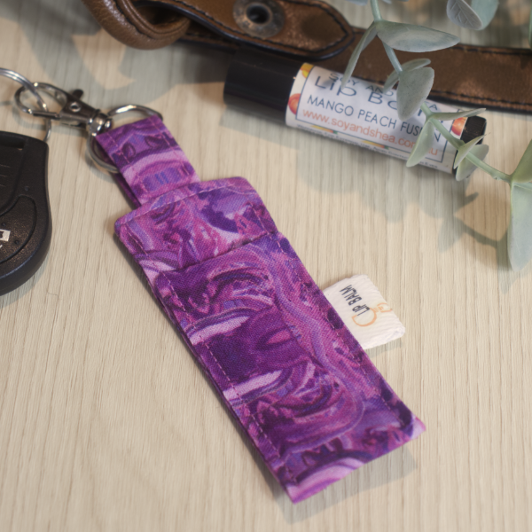 A clip balm holder lies on a wooden surface attached to a car key with a "lobster claw" clip.  To the side is a tube of lip balm surrounded by greenery and a leather strap from a bag.  The Clip Balm has a rich pink and purple swirl pattern