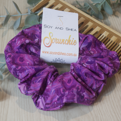 A colorful scrunchie sits on wooden surface resting against a wooden comb with greenery in the background.  A cardboard tag reads SOY AND SHEA SCRUNCHIE.    The fabric features a tie dye pattern in shades of bright to dark purple