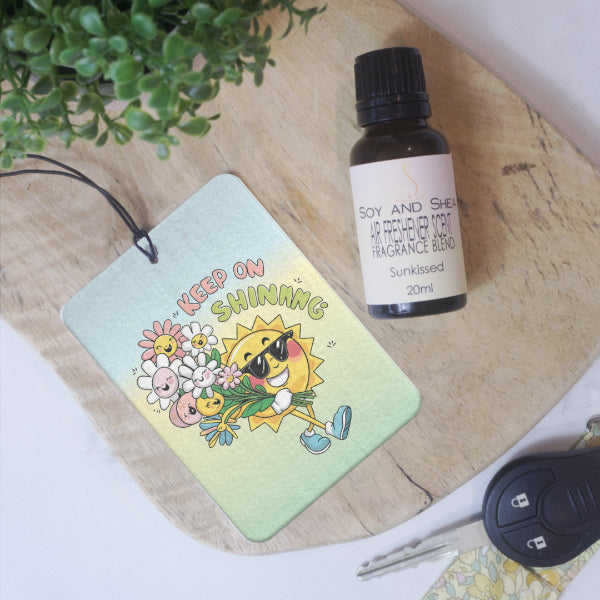 A rectangular car air freshener lies on a wooden surface next to a glass bottle of Air Freshener Scent and car key.  The Air Freshener features a drawing of a smiling sun with legs carrying a bunch of pink and white flowers all with smiling faces on a ombre background of  blue to green.  Text reads KEEP ON SHINING