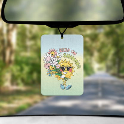 A mock up image showing a view through a car windscreen with a long tree lined road.  From the rearview mirror hangs an air freshener that has an ombre background of blue at the top green towards the bottom.  It has a cartoon image of a smiling sun walking along with a bunch of flowers all with smiling faces next to text reading KEEP ON SHINING